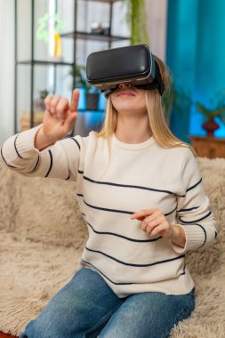Beyaz kadın evde kanepenin üzerinde sanal gerçeklik oyunu oynarken VR kulaklığı takıyor. Kız heyecanlı, şaşırmış, aktif, kolları hızlı hareket eden, dijital ortama dalmış, şaşkın ve neşeli görünüyor.