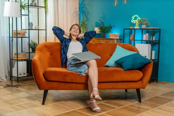 Oturma odasındaki işi bittikten sonra kanepeye oturmuş laptopu kapatan beyaz bir kadın. Mutlu genç kız serbest çalışıyor. Ev kanepesinde uzaktan kumandayla çalışıyor. Defter bilgisayarında e-öğrenme, internette gezinme