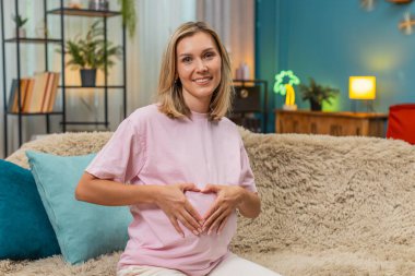 Hamile, orta yaşlı bir kadın karnının üzerinde elleri ile kalp oluşturur. Yavaşça gülümser ve doğum için beklenti hisseder. Kafkasyalı kız kanepeye uzanmış, nazik bir anın tadını çıkarıyor. Yakında bebeğe sessiz bir aşk yolluyor..