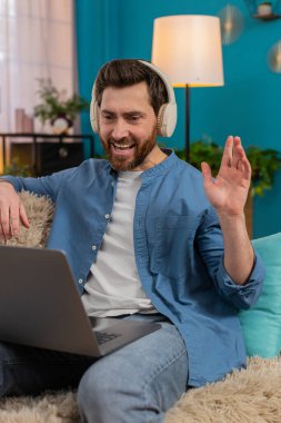 Evde orta yaşlı bir adam, kablosuz kulaklık takıyor, webcam 'e el sallıyor ve videonun keyfini çıkarırken gülüyor. Kanepede oturan beyaz adam, arkadaşlarıyla veya meslektaşlarıyla sohbet etmek için dizüstü bilgisayar kullanıyor.