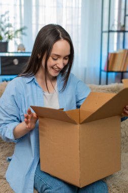 Genç, mutlu bir kadın, mutlu bir şekilde kutuları açıyor. Arzu edilen eşyaları çıkarırken hoş bir sürpriz yaşıyor. Kanepede heyecanlı beyaz bir kız, online alışveriş paketinden neşeli duygular.