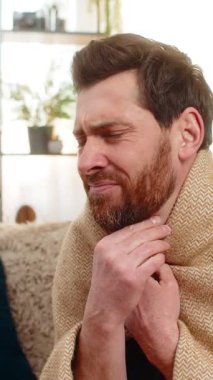 Evdeki genç adam kanepeye oturur, battaniyeye sarınır, öksürür ve acı içinde boğazını tutar. Kafkasyalı adam kaşlarını çatmış, yutkunmaya çalışmış, muhtemelen boğaz ağrısı ya da grip belirtileri göstermiş.