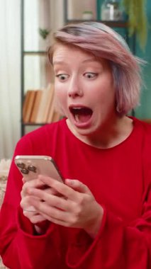 Evdeki akıllı telefon ekranına şaşırmış, beklenmedik para transferine şaşırmış genç bir kadın. Kafkasyalı kız heyecanla haykırıyordu. Mutlu ve şaşkın bir yüz ifadesiyle koltukta oturuyordu..