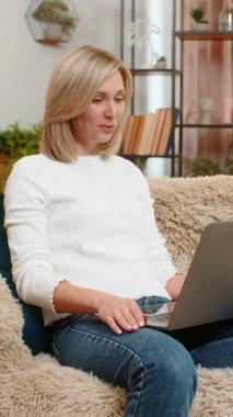 Orta yaşlı bir kadın video toplantısı sırasında evdeki kanepede konuşuyor, gülümsüyor ve anlamlı merhaba jestleri yapıyor. Yetişkin kız, pozitif enerji ve çatışma göstergesi olarak çevrimiçi ekiple güven içinde iletişim kuruyor..