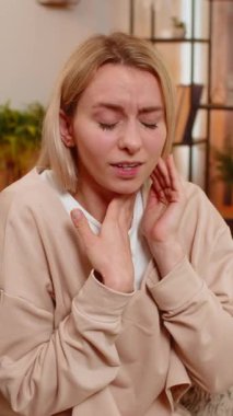 Genç yetişkin bir kadın evdeki koltukta boğazına dokunuyor, yutkunmaya çalışırken açıkça acı gösteriyor. Sarışın kız gözle görülür bir rahatsızlıkla tepki veriyor. Boğaz bölgesinde iltihaplanma ya da hastalık belirtileri gösteriyor..