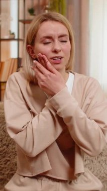 Genç yetişkin bir kadın koltukta otururken güçlü diş ağrısı belirtileri göstererek acı içinde yanaklarını tutuyor. Sarışın kız kaşlarını çatar ve rahatsızlık gösterir, diş sorunu ve hassasiyete duygusal tepki verir.