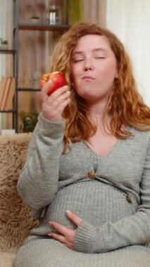 Hamile, genç, mutlu bir kadın bir eliyle karnına hafifçe dokunarak kanepeye oturur, taze bir elma ısırır, besleyici atıştırmalıkların tadını çıkarır. Kız huzur ve tatmin hissediyor, sağlıklı seçimleri kucaklıyor.