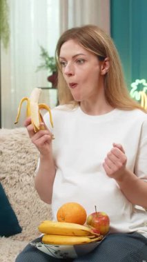 Evde orta yaşlı hamile bir kadın meyveli atıştırmalık yemekten muzlu gösterilerden zevk alıyor. Sağlığa önem veriyor. Kanepede oturan beyaz bir kız elma tabağıyla gülümsüyor vitaminleri seviyor bu yüzden günlük diyet bebeği güçlü tutuyor..