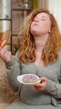 Hamile, genç, mutlu bir kadın elinde donutla kanepede oturuyor. Neşeli bir ifadeyle gülüyor, tatlı bir zevkin tadını çıkarıyor. Kız küçük zevkleri kucaklıyor, mutluluğu basit ve lezzetli anlarda buluyor..