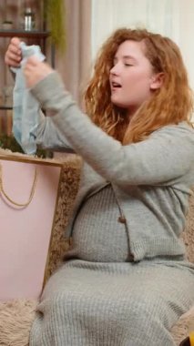 Hamile genç bir kadın alışveriş çantalarını evde bebek kıyafetleriyle açıyor, heyecandan ve memnuniyetten gülümsüyor. Kız, gelecekteki anneliğe hazırlanmaktan hoşlanıyor. Küçük dönüm noktalarını olumlu bir şekilde kutluyor.