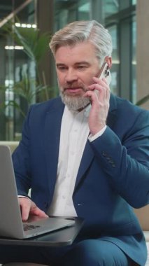 Kendine güvenen, takım elbiseli, akıllı telefon ve dizüstü bilgisayar kullanarak müşteriye cevap veren olgun bir iş adamı. Sohbet. İnternette çalışan beyaz sakallı adam modern ofis lobisinde konuşuyor..
