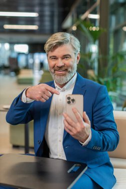 İnternette akıllı telefon uygulaması kullanarak gülümseyen yetişkin işadamı ofis lobisinde çalışan uzaktan kumandayı işaret ediyor. Takım elbiseli adam sosyal medyada e- posta içeriği öğreniyor.