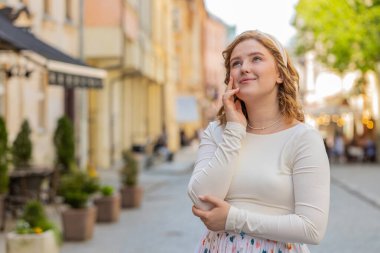 Düşünceli, güler yüzlü, uzun elbiseli, düşünceli, mutlu, genç bir kadın. Düşünceli bir yüz ifadesi var. Çözüm düşünüyor. Şehir caddesinde yürüyen kız.