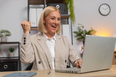 İç işlerinde masa başında otururken laptoptan harika haberler okuyan mutlu beyaz kadın başarısını kutluyor. Gülümseyen iş kadını güzel bir tebliğden memnun, evet işareti gösteriyor..