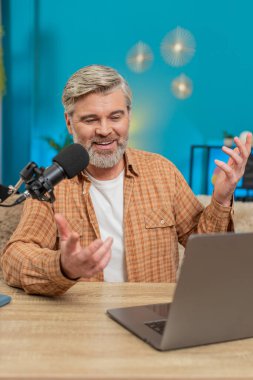 Olgun adam blogcusu canlı yayın akışı vlog 'u sırasında izleyicilerle etkileşim halinde olan stüdyo mikrofonuna konuşuyor. Orta yaşlı bir adam evde oturmuş jestler yapıyor, geri bildirimler yapıyor. Dikey