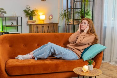 Hasta kızıl saçlı kadın baş ağrısı problemini tedavi etmek için tapınakları ovuyor, tansiyon, migren, stres, ekşitme, evde yüksek tansiyon. Genç yetişkin bir kız odada kanepede..