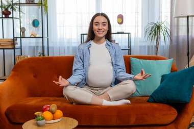 Oturma odasında rahat bir kanepede meditasyon yaparken nilüfer pozisyonunda oturan hamile, çekici bir kadın. Sağlık, hamilelik ve nefes alma pratiği kavramı. Gülümseyen bayan anne olmaya hazırlanıyor.