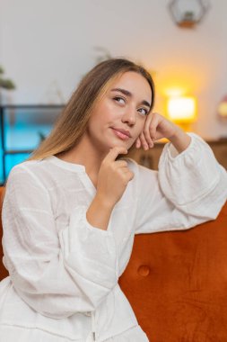 Beyaz elbiseli, düşünceli, beyaz tenli, çenesine dokunarak hafta sonu için nereye gideceğini düşünen, evdeki oturma odasındaki kanepede oturan yeni fikirler üreten genç, çekici beyaz bir kadın. Dikey