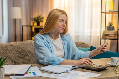 Kadın evde harcamaları düzenler, rakamları not defterine yazar ve faturaları sabırla onaylar. Masadaki beyaz kız, finans için berraklık ve dikkatli strateji birleştirme hesap makinesi kullanıyor