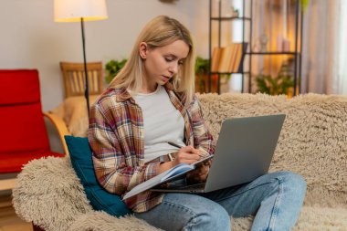 Genç yetişkin bir kadın dizüstü bilgisayar ekranına odaklanırken deftere yazar. Masrafları hesaplar. Sarışın kız dikkatli bir şekilde mali kayıtları yönetiyor, düzenli ve işine odaklı kalıyor..