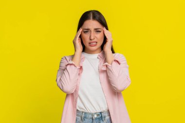 Güçlü baş ağrısı ve rahatsızlıktan muzdarip acı dolu bir ifadeyle iç çekip el ele tutuşan genç bir kadın. Sarı arka plandaki kız açıkça gerginlik, yorgunluk ve duygusal gerginlik gösteriyor.