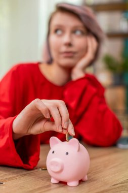 Genç bir kadın kumbaraya madeni para koymaktan mutlu. Ev masasında oturmuş, mali disiplinle gülümsüyor. Beyaz kız dikkatli para biriktirme alışkanlıkları sergiliyor, gelecekteki hedefler için akıllıca bütçe ayırıyor..