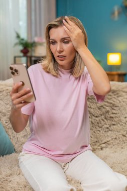 Yenilgide online oyun omuzlarını kaybettikten sonra evde oturup akıllı telefon kullanan hamile bir orta yaşlı kadın. Kanepedeki beyaz kız üzgün yüzünü gösteriyor. Titreyen kafa cep telefonu ekranına bakıyor ve üzgün hissediyor..