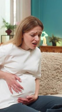 Hamile orta yaşlı kadın evde karnını sıkıyor hafif kasılmalar sırasında nefes alıyor sonra rahatlıyor ve küçük tekmeler hissederek gülüyor. Kanepede oturan beyaz kız bebek hareketlerini neşeyle ve sakin bir şekilde dinliyor..