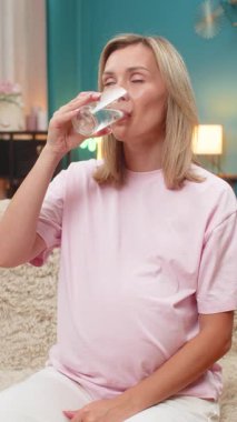 Hamile, orta yaşlı bir kadın karnına su dökerek hamilelik boyunca susuzluğun önemini gösterir. Kanepedeki beyaz kız sıvı alışkanlığıyla bebek sağlığını koruduktan sonra serinledi..