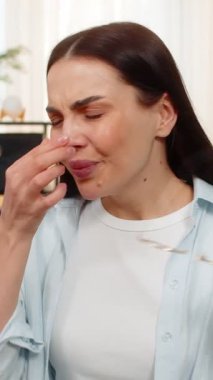 Evde genç bir kadın göz ağrısı ve rahatsızlık hissediyor. Gözlüklerini çıkartıyor ve gözlerini nazikçe ovuyor. Kanepede oturan beyaz hasta kız görsel gerginlik yaşıyor, ekran çalışmasından yorgun düşmüş ya da kitap okuyor.