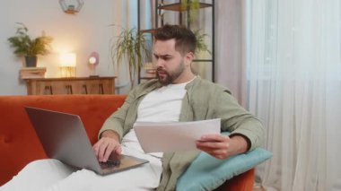 Kanepede dizüstü bilgisayarını kullanarak evrak işleriyle uğraşan genç beyaz bir adam. Gayri resmi kıyafetleri olan üzgün bir adam. Evrakları hava kapama ağlarına atıyor ve evdeki oturma odasına bakıyor..