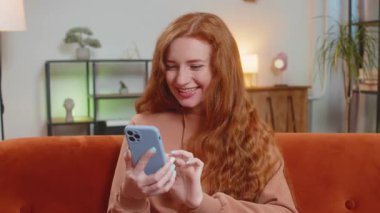 Evdeki oturma odasında gülümseyen cep telefonu kullanan mutlu, genç, kızıl saçlı bir kadın. Kızıl Kız akıllı telefon sosyal medya uygulamalarında internetten film izlerken mesajlarını paylaşıyor.