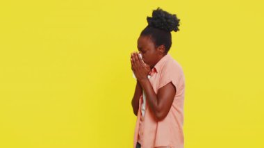 Afro-Amerikalı kadın hapşırır ve burnu peçeteyle siler. Yorgunluk hissi verir. Sarı arka plandaki siyah kız soğuk, burnu akan, yorgunluk, virüs ve alerji belirtileri gösteriyor..