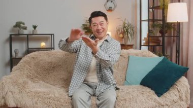Cheerful happy rich Asian man showing wasting, throwing money around hand gesture, more tips earnings, big profit, win lottery, body language, share, celebrate. Guy at home in living room apartment.