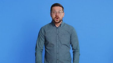 Kafasına dokunup kafa patlaması gösteren, endişeli ve şok olmuş, profesyonel tükenmiş, gözlüklü, orta yaşlı, heyecanlı bir beyaz adam. Genç yetişkin adam mavi arka planda izole edilmiş