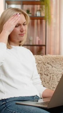 Orta yaşlı bir kadın dizüstü bilgisayar kullandıktan sonra kafasına masaj yaparak yorgun ve stresli bir şekilde evde oturuyor. Yetişkin kadın, baş ağrısı ve gerilimden kurtulmak için aşırı çalışma ve dijital baskıdan dolayı acı hissediyor.