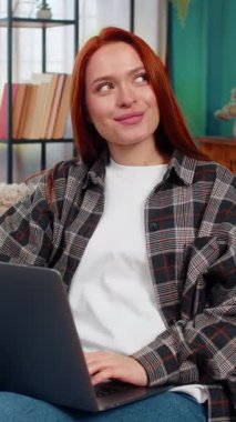 Genç bir kadın dizüstü bilgisayarında internetten elbise seçiyor ve düşünürken çenesine dokunuyor. Kanepedeki beyaz saçlı kız alışveriş kararına odaklanmış, moda ve dijital alışverişin tadını çıkarıyor.