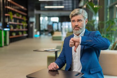 Hoşlanmamak. Takım elbiseli üzgün işadamı ofis lobisinde oturmuş işaret dilini gösteriyor hoşnutsuzluğunu, memnuniyetsizliğini ve kötü işini dile getiriyor. Otel odasında ciddi bir olgun adam var..