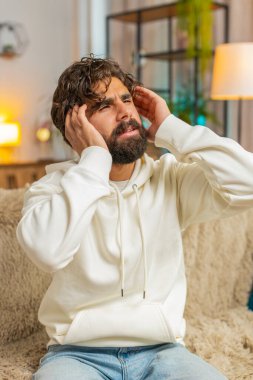 Kapüşonlu genç adam baş ağrısı problemini tedavi etmek için tapınağı ovuyor, tansiyon ve migrenden muzdarip, evde acı içinde kıvranıyor. Kıvırcık saçlı genç adam kanepede oturuyor..