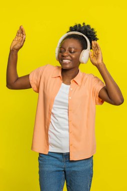 Neşeli Afrikalı Amerikalı kadın kulaklıkla müzik dinliyor, dans ediyor, eğleniyor, el kol hareketleri yapıyor. İçerideki sarı arka planda izole edilmiş genç siyah kız.