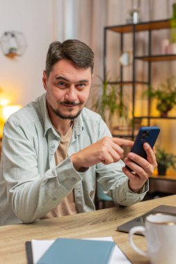 Evde gülümseyen cep telefonuyla masada oturan mutlu beyaz bir adam. Serbest çalışan biri, internetten akıllı telefon sosyal medya uygulamalarından mesaj yolluyor. Dikey.