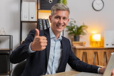 Gibi. Kafkasyalı mutlu işadamı onaylayıcı bir şekilde kameraya bakıyor ve olumlu bir işaret veriyor iyi haber olumlu geri bildirim. Genç yakışıklı adam evdeki masada oturuyor..