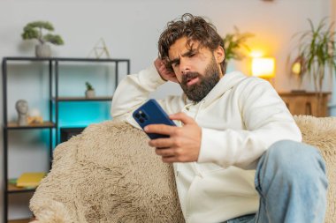 Oturma odasındaki kanepede oturan akıllı telefon kullanan mutsuz Hintli adam. Kafasına dokunan kapüşonlu genç İspanyol adam aniden piyango ya da kötü haber karşısında şaşırdı..