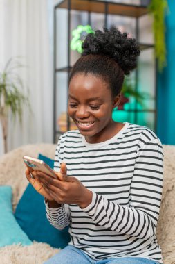 Afrikalı Amerikalı kadın sosyal medya profilini güncelliyor. Evdeki kanepede oturmak, fotoğraf çekmek, tasvirler yazmak, mime gülmek. Rahatlamış ruh hali, kapalı mekanda gelişigüzel etkileşim. Çevrimiçi zaman geçirmek eğlenceli