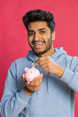 Kumbarayı elinde tutan mutlu Hintli genç adam bozuk para bırakarak para tasarrufu yapıyor, gelecekteki harcamaları ve finansal güvenliği planlıyor. Kırmızı arka planda izole edilmiş, tasarrufa yatırım yapan gülümseyen Arap adam.