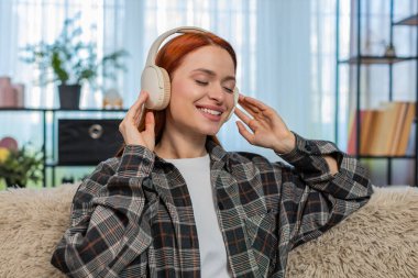 Evde kablosuz kulaklık takan, gözleri kapalı, favori müzik dinleyen genç bir kadın. Kanepede oturan beyaz saçlı kız sakin duygusal dengesiyle huzurlu bir ses anının tadını çıkarıyor..