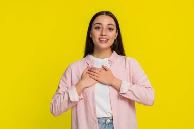 İki elini de göğsüne kaldırmış, kaşlarını kaldırmış genç bir kadın minnettarlığını açıkça ifade ediyor. Sarı arka plandaki kız samimi duygular ve içten şükran dolu bir tepki gösteriyor.