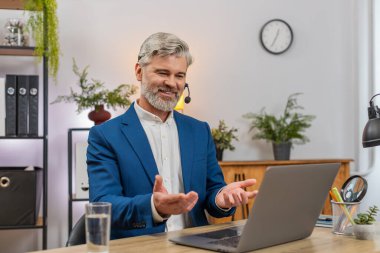 Merkez ofisteki orta yaşlı gülen işadamı laptopla yardım hattını arayan kişiye kulaklık takıyor. Masadaki serbest çalışan müşteri memnuniyetini artırmak için hızlı destek veriyor.