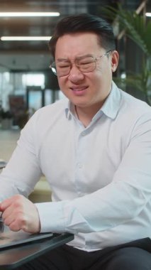 Gergin Çinli yetişkin iş adamı dizüstü bilgisayar kullanıyor ve modern ofis lobisinde çalışırken kalp krizi geçirme riski olan bir göğsü tutmaktan muzdarip. İş adamı gömlekli..