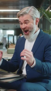 Dizüstü bilgisayarda çalışan beyaz sakallı iş adamı ani zafer, piyango hedefi başarısı ve iyi bir kariyer haberiyle şok oldu. Modern otel lobisinde oturan heyecanlı adam yumruklarını sıkıyor..